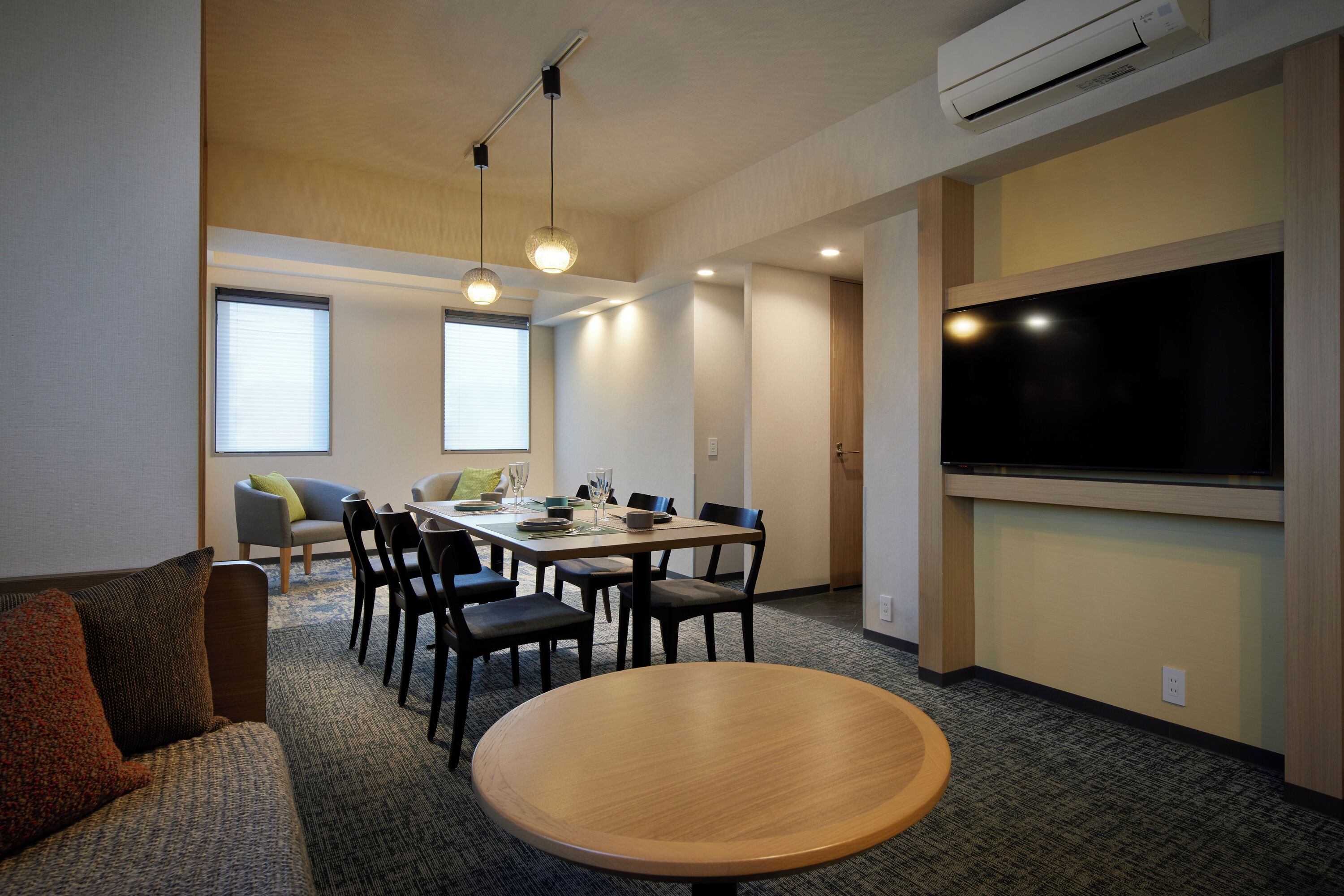 A conference room with a round table, rectangular table, and chairs. There is a TV on the wall and an air conditioning unit above the table.