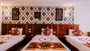 A hotel room with three beds, each with a white bedspread and red and gold decorative pillows. The room features a patterned wallpaper and wooden headboard.