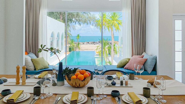 A dining area with a view of the beach and ocean, featuring a pool, seating with cushions, and a table set for a meal. - Cape by Sichon Tesoro (Sichon)