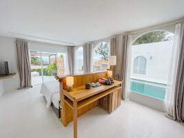 A hotel room with a wooden desk, a bed, a TV, and a view of a swimming pool.