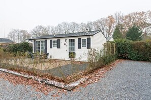 A white building with black shutters, a gravel driveway, and a small garden area with leaf-covered ground.