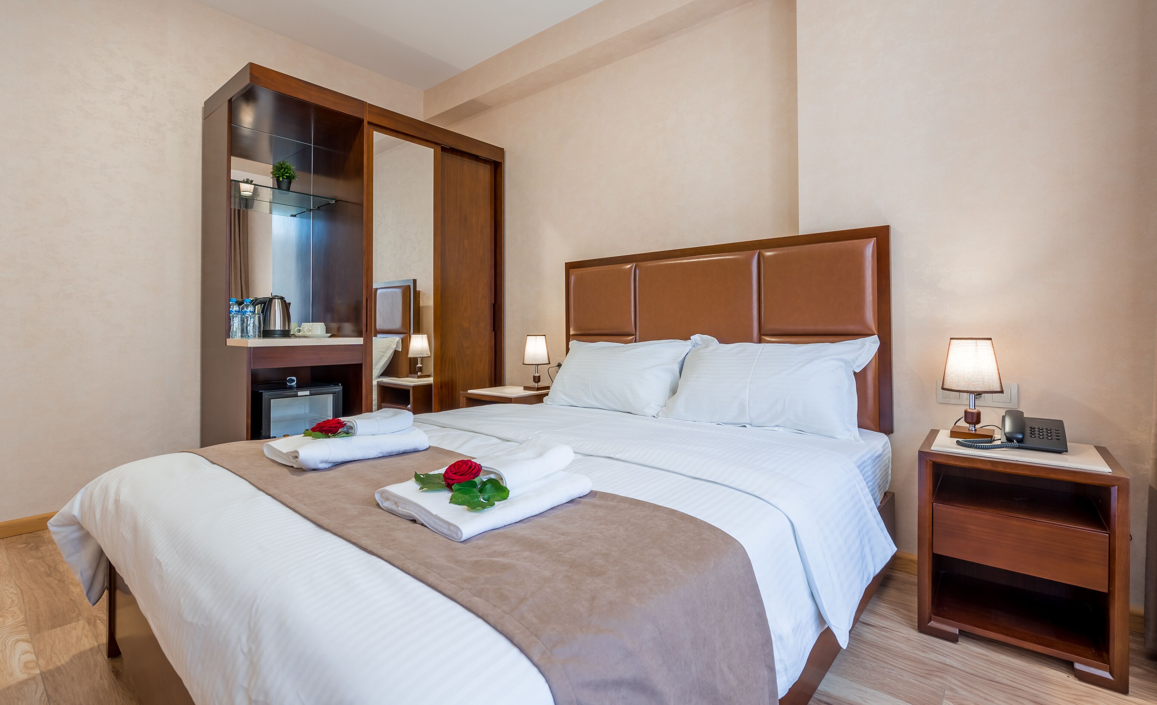 Habitación de hotel con una cama grande, cabecero de madera, dos mesitas de noche con lámparas, un espejo, un escritorio pequeño con una silla y un estante con artículos de aseo.
