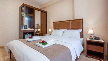 Habitación de hotel con una cama grande, cabecero de madera, dos mesitas de noche con lámparas, un espejo, un escritorio pequeño con una silla y un estante con artículos de aseo.