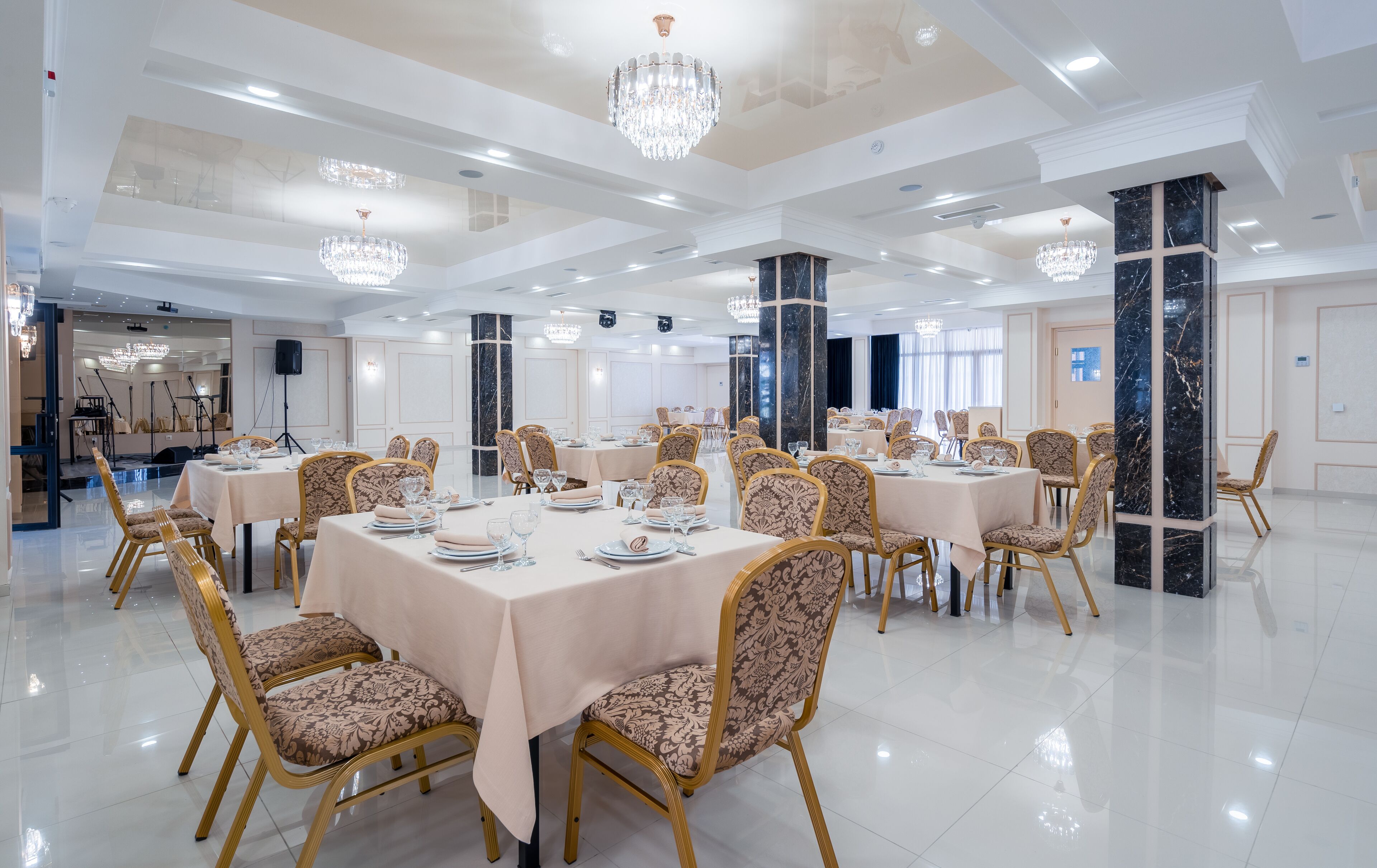 A spacious banquet hall with multiple round tables, elegant chandeliers, and marble columns.