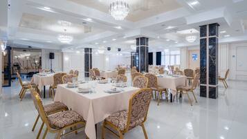 A spacious banquet hall with multiple round tables, elegant chandeliers, and marble columns.