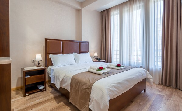 A hotel room with a large bed, a nightstand, a wooden shelf, and a window with curtains.