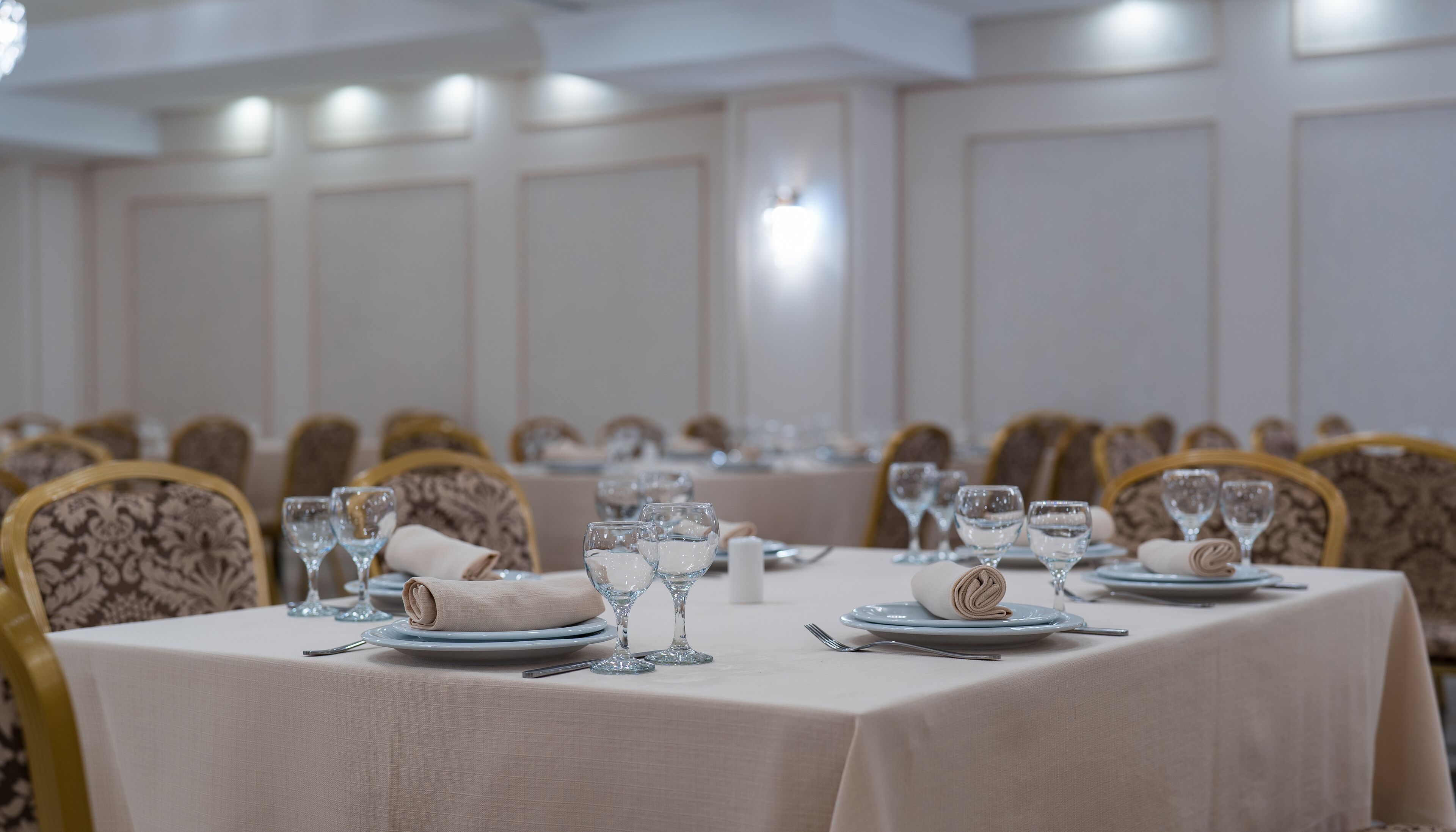 Une salle de banquet avec des tables dressées pour le repas, des chaises recouvertes d’un tissu à motifs et une palette de couleurs neutres.