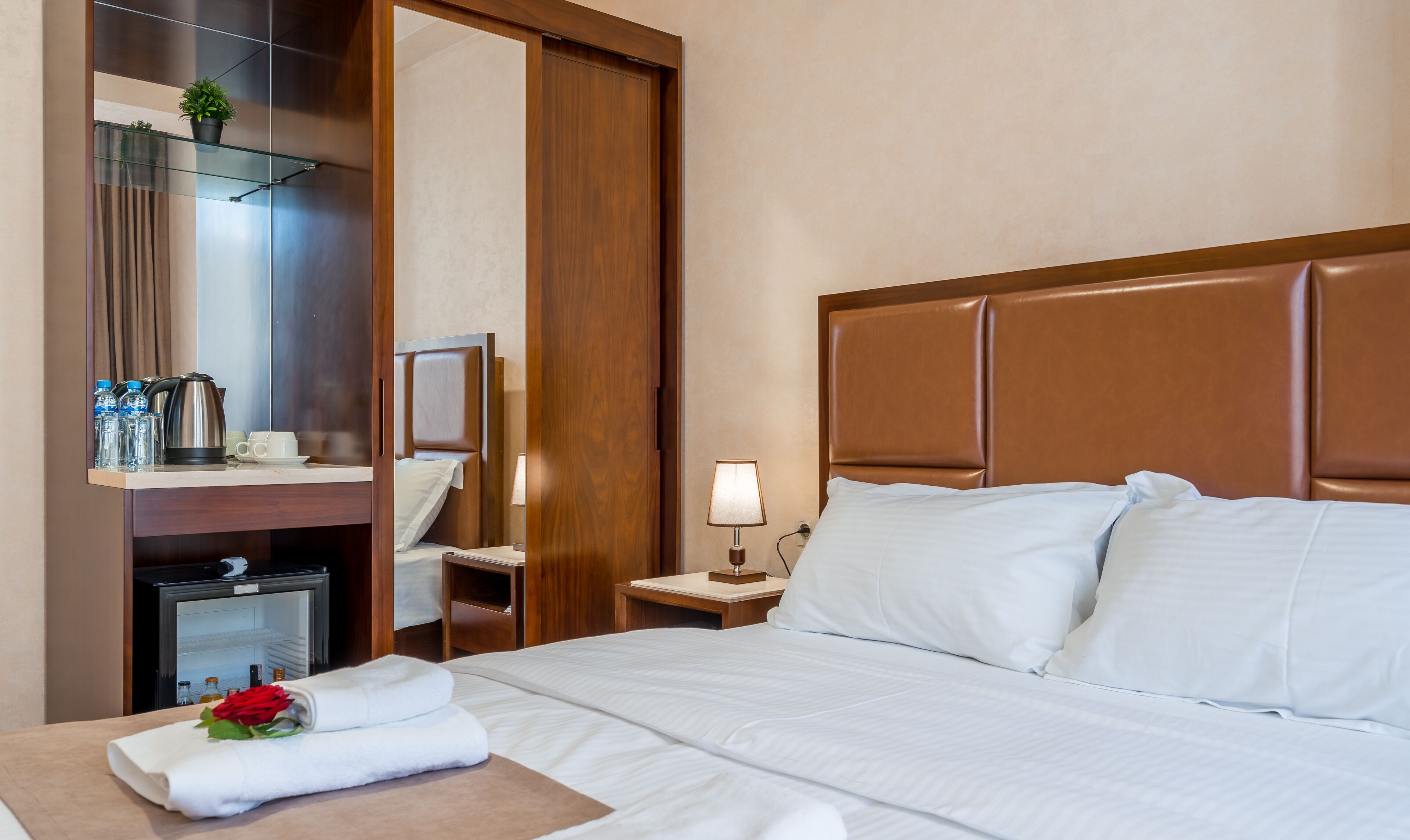 Een hotelkamer met een bed, nachtkastje met lamp, houten hoofdbord, spiegel, minibar en een kleine kamerplant.
