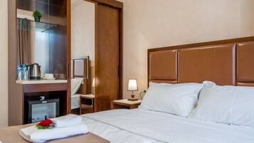 A hotel room with a bed, a nightstand with a lamp, a wooden headboard, a mirror, a minibar, and a small potted plant.