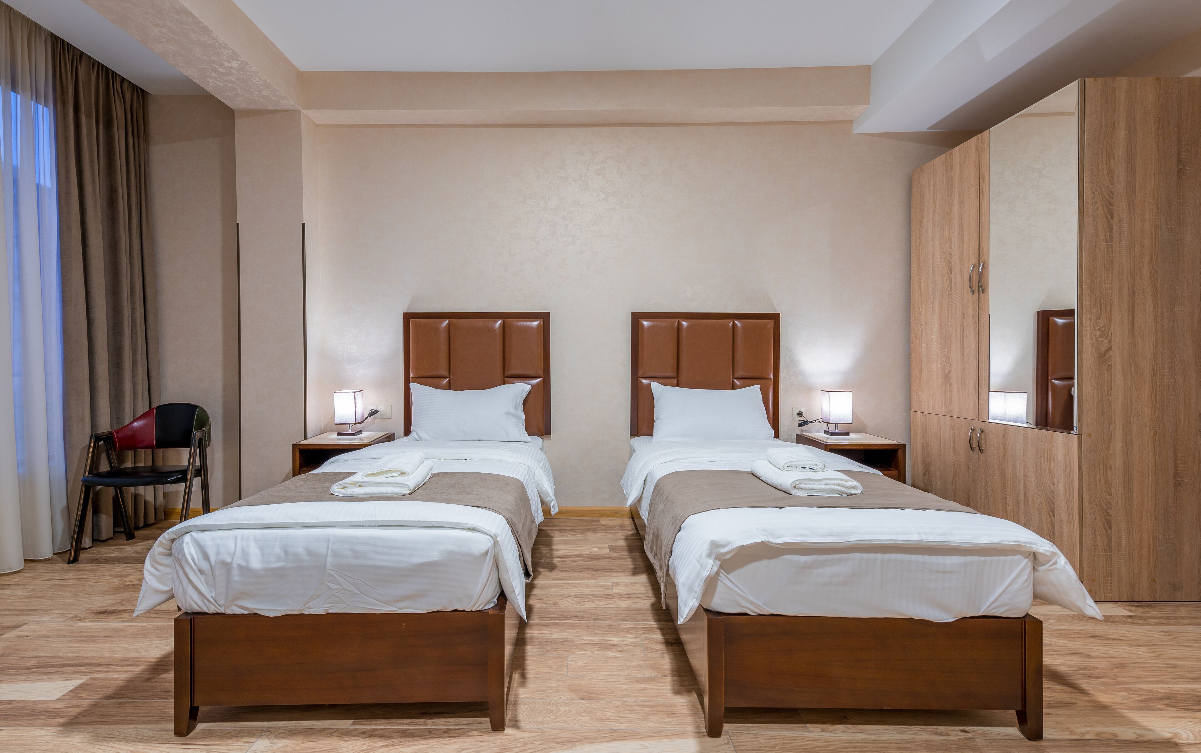 Una habitación de hotel con dos camas, cabeceros de madera, mesitas de noche con lámparas, una silla y un espejo grande.
