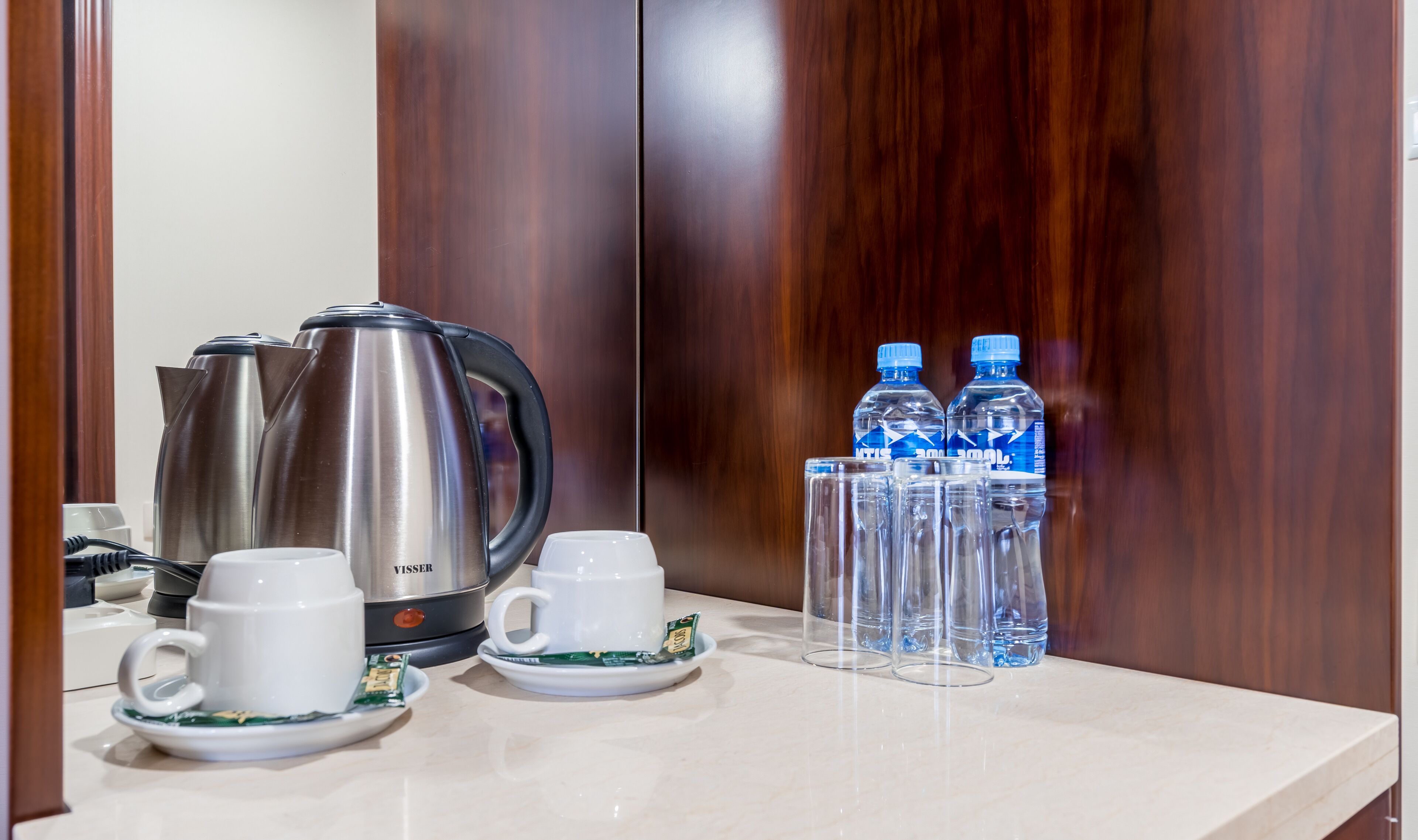 Una kitchenette con un hervidor de acero inoxidable, dos tazas blancas con platillos y dos botellas de agua sobre una encimera de mármol.