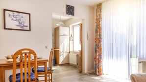 Un coin repas avec une table et des chaises en bois, une horloge au mur et une vue sur la cuisine par une porte ouverte.