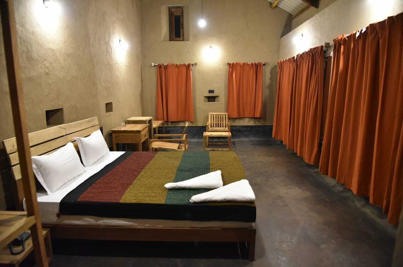Habitación individual con una cama de madera, mesita de noche, una silla y una pequeña mesa. Cortinas anaranjadas cubren las ventanas.
