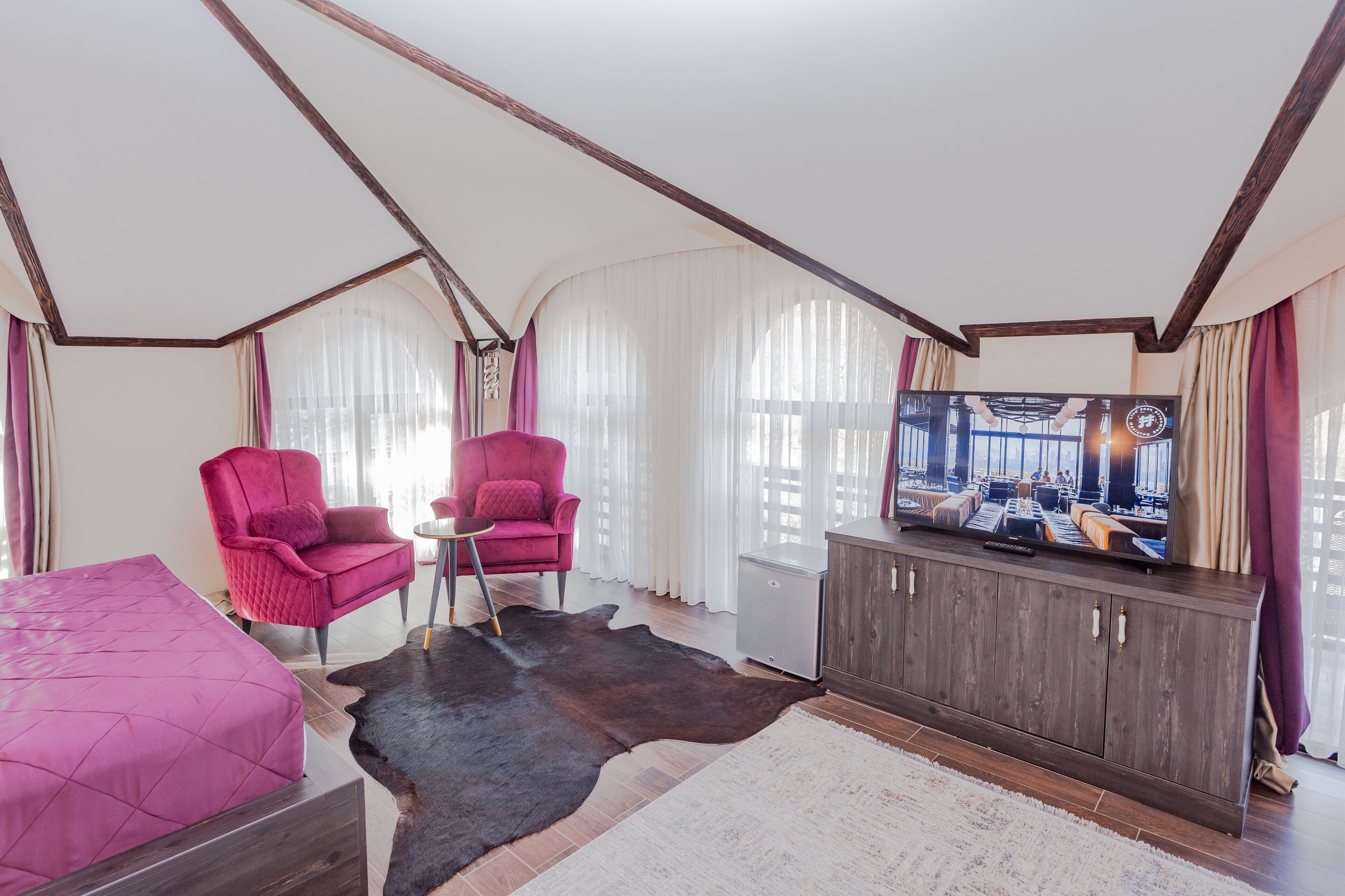A room with a sloped ceiling, a large flat-screen TV, a wooden cabinet, two pink armchairs, a bed with a pink bedspread, and a cowhide rug.
