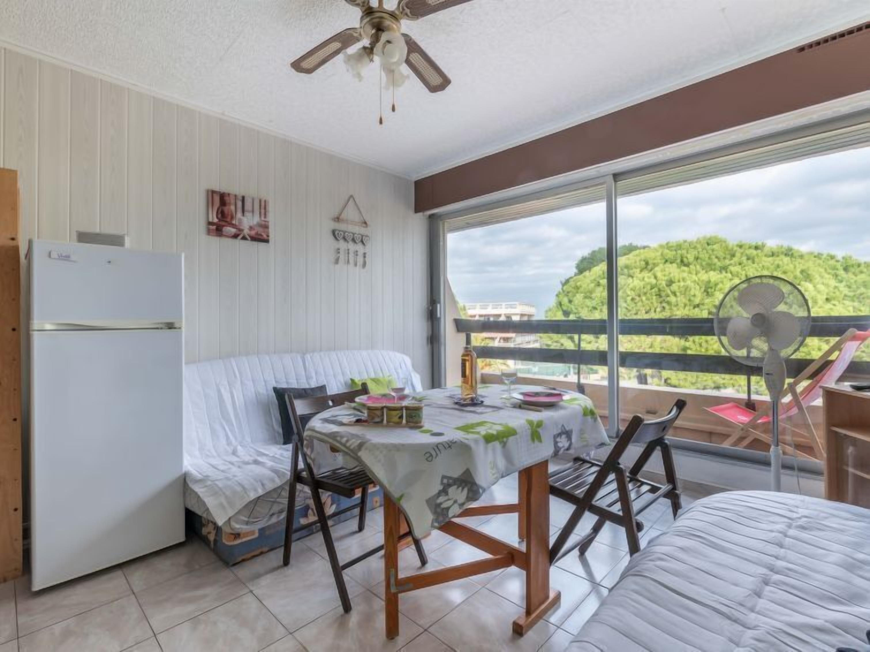 Habitación compacta con cama, una mesita con mantel, ventilador de techo, refrigerador y balcón con vista a la vegetación.