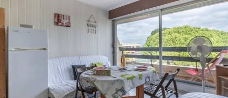 Habitación compacta con cama, una mesita con mantel, ventilador de techo, refrigerador y balcón con vista a la vegetación.