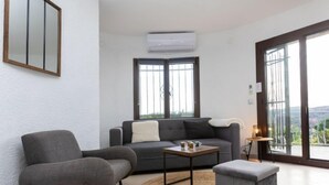 Un soggiorno moderno con un divano grigio, un tavolino, una poltrona grigia e un piccolo poggiapiedi. Si vede una grande finestra con vista sull'esterno, un condizionatore d'aria a parete e uno specchio sopra il divano.
