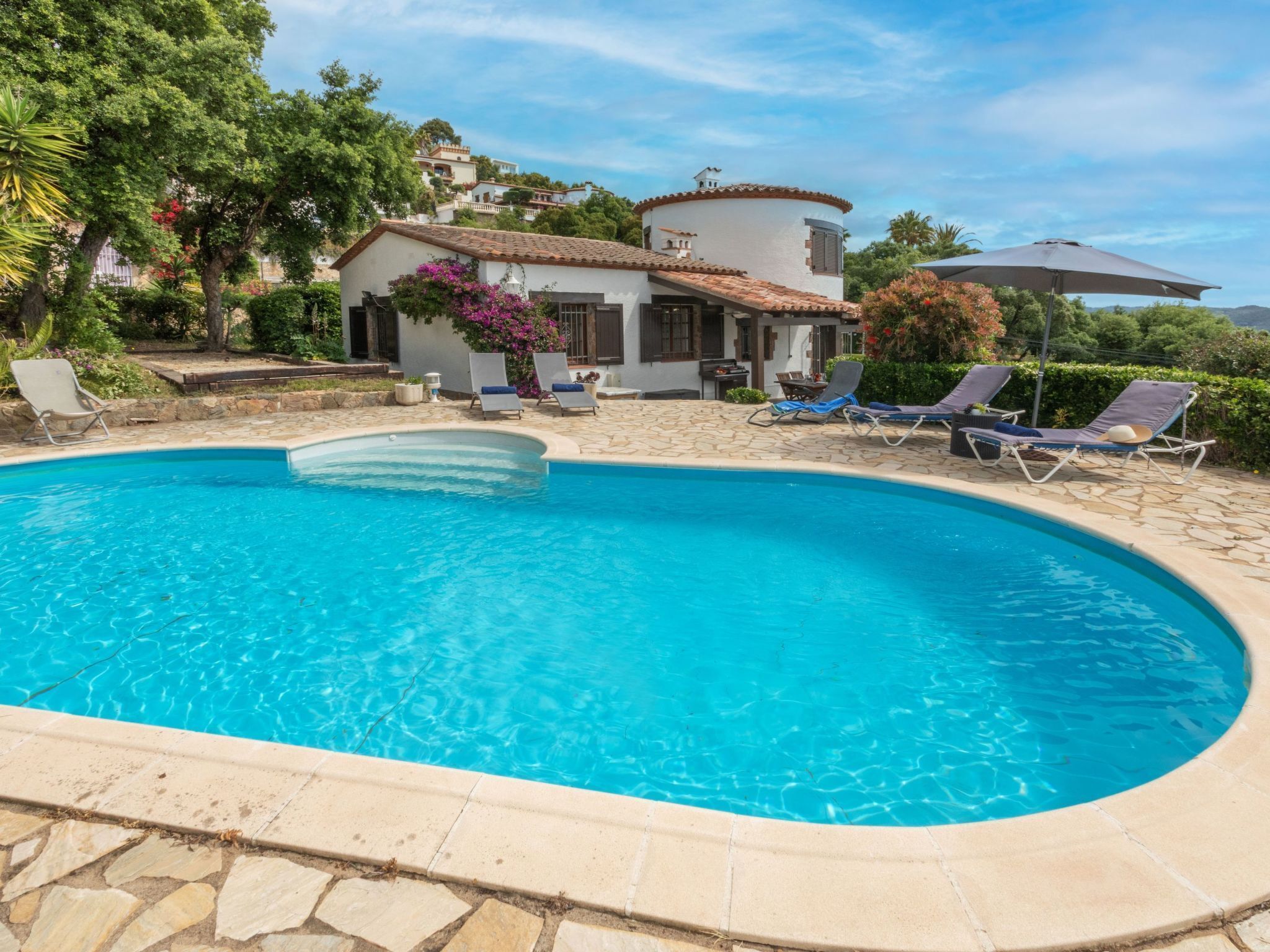 En swimmingpool med liggestole, en stenbelagt terrasse og et hus med et tag i fliser.