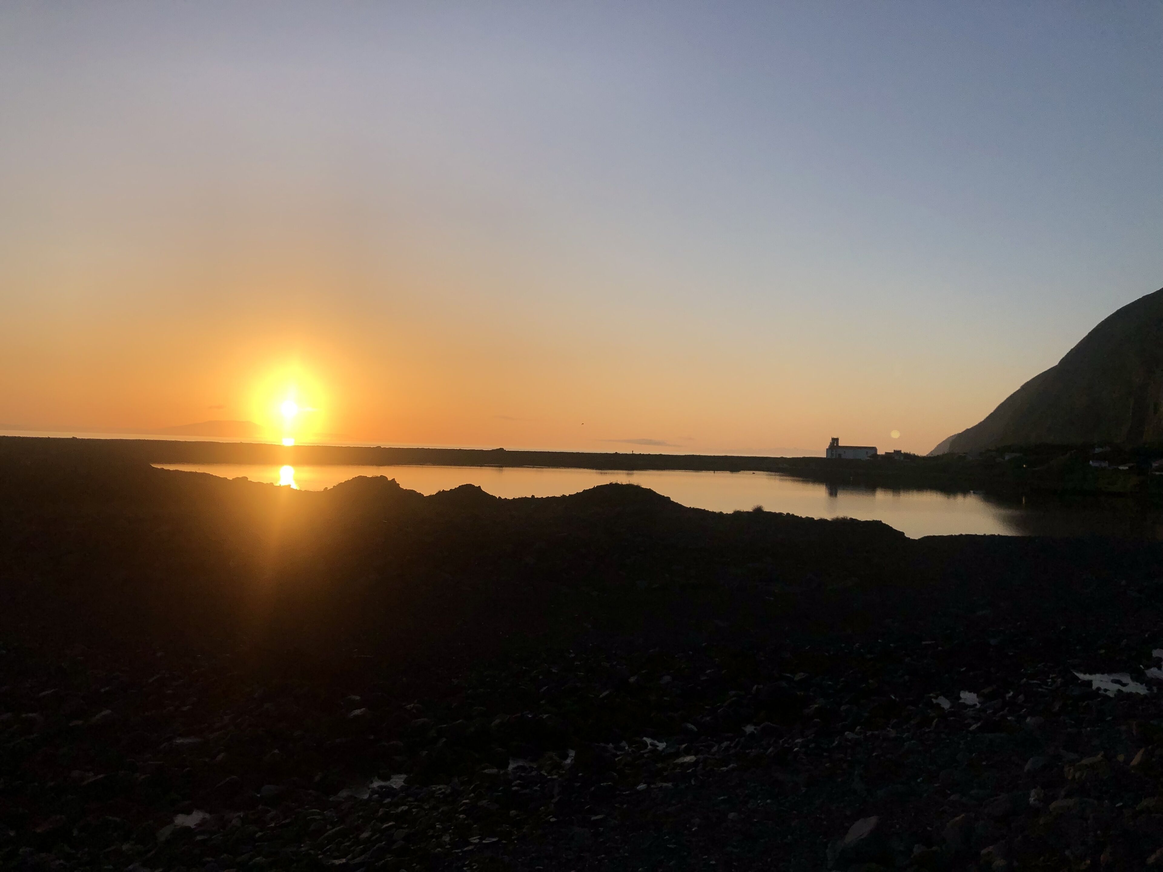 Um pôr do sol sobre águas calmas, com uma costa rochosa e um edifício ao longe.