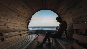 Eine Person sitzt auf einer Holzbank in einer Holzhütte mit Blick auf das Meer.