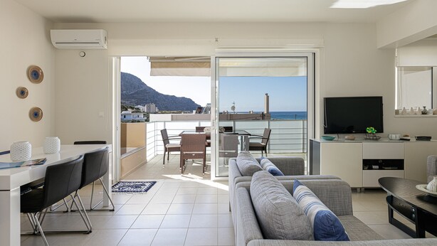 Ein modernes Wohnzimmer mit einer Couch, einem Esstisch und Stühlen, einem Fernseher und einem Balkon mit Blick auf das Meer und die Berge.