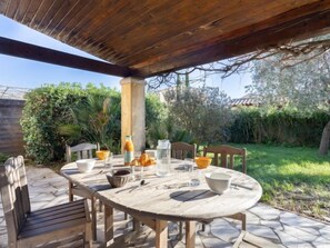 Une table ronde en bois, entourée de chaises, se trouve sous une pergola en bois, elle-même bordée de verdure et d’un chemin en pierre.
