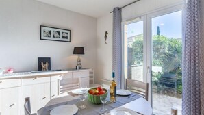 Een eetgedeelte met een tafel voor twee, een schaal fruit en een fles wijn. Er is een raam met gordijnen, een lamp en een ingelijste foto aan de muur.