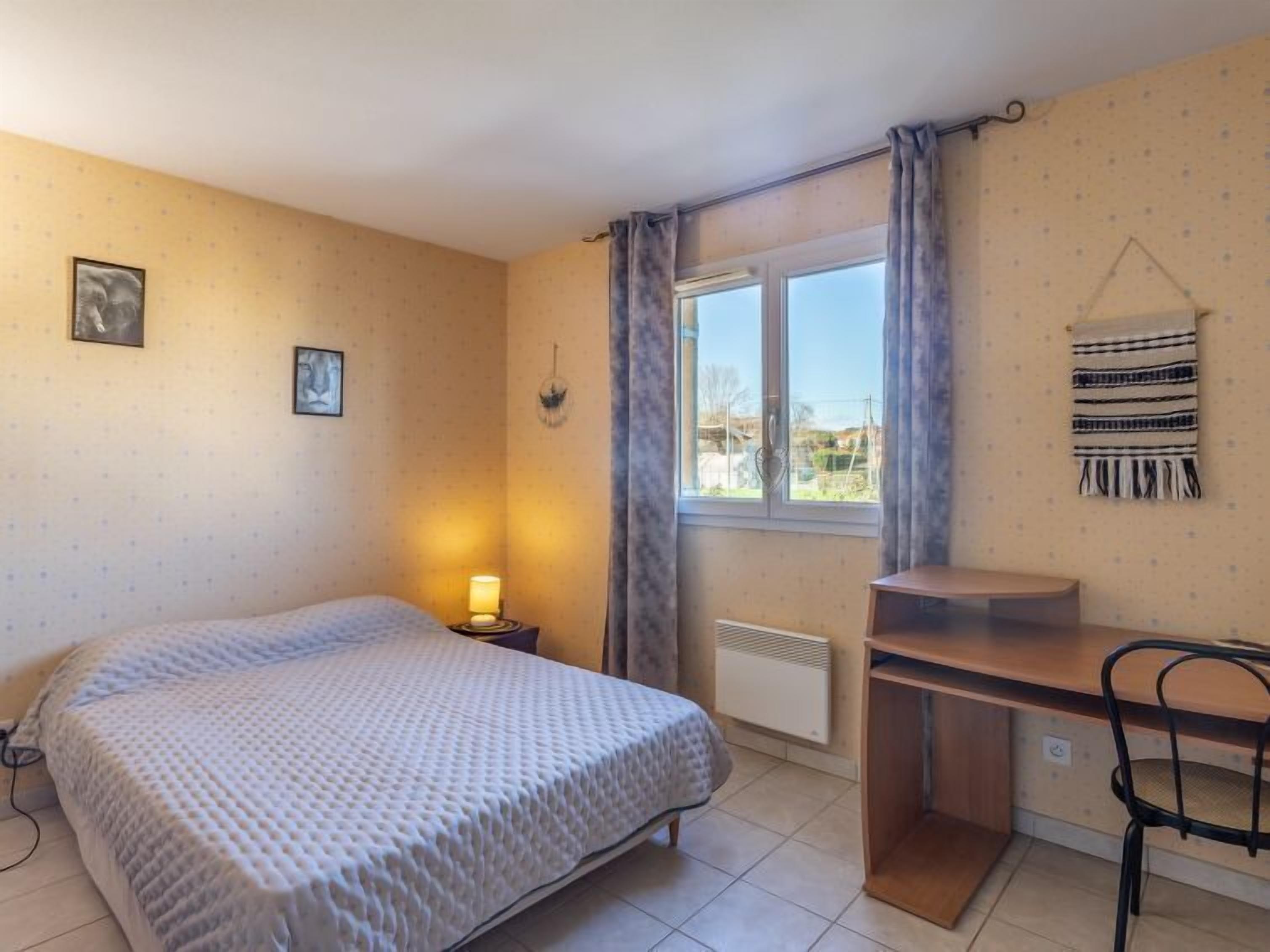 Quarto com cama, escrivaninha, cadeira, janela com cortinas e radiador.