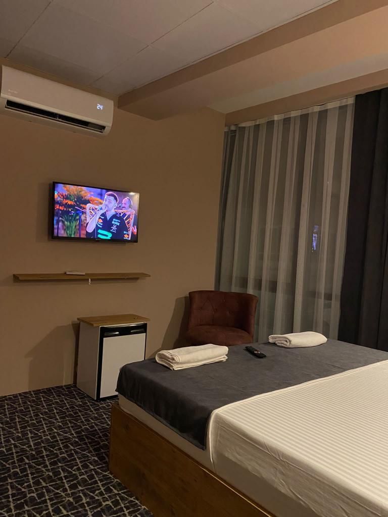 Habitación de hotel con cama, televisor montado en la pared, una silla, una mesita y una ventana con cortinas.