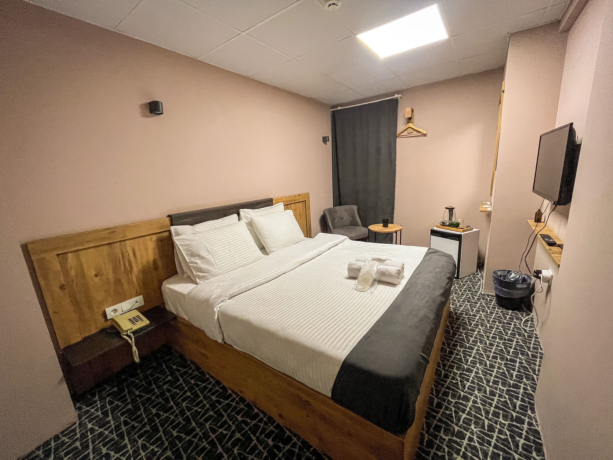 Une chambre d’hôtel avec une tête de lit en bois, un lit recouvert de draps blancs, un fauteuil gris, une petite table avec une lampe et un téléviseur fixé au mur.