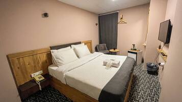 Une chambre d’hôtel avec une tête de lit en bois, un lit recouvert de draps blancs, un fauteuil gris, une petite table avec une lampe et un téléviseur fixé au mur.