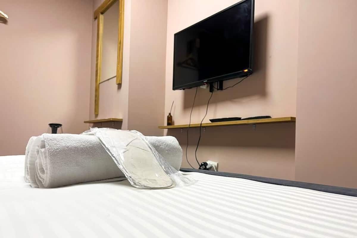 Una cama bien hecha con una toalla blanca enrollada y una bolsa de plástico, un televisor de pantalla plana montado en la pared y un estante de madera debajo.