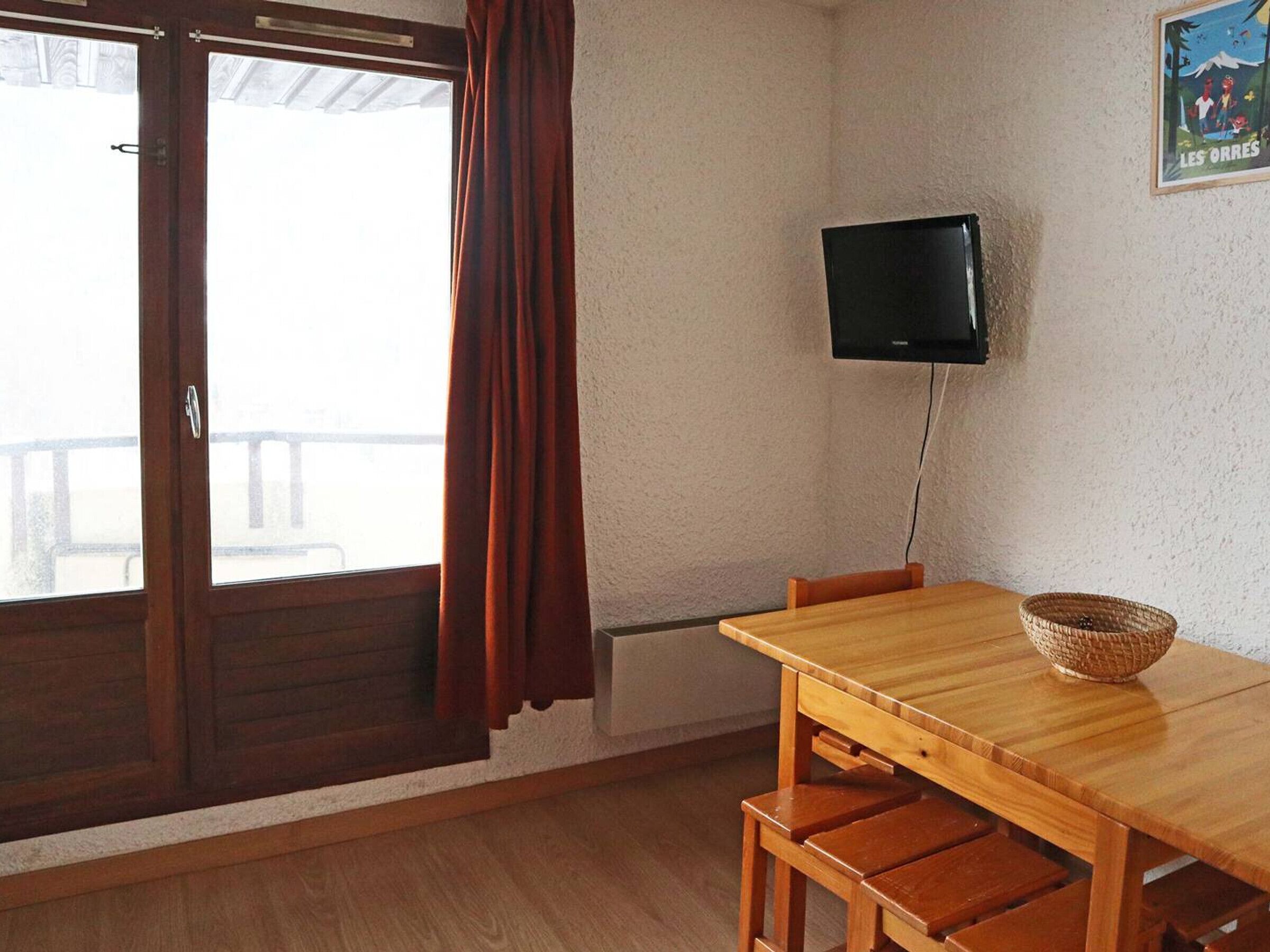 A room with a wooden table and chairs, a television mounted on the wall, and a window with curtains.