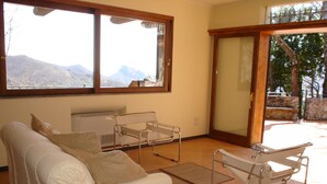 A room with a white sofa, a metal-framed chair, a radiator, and a view of mountains through large windows.