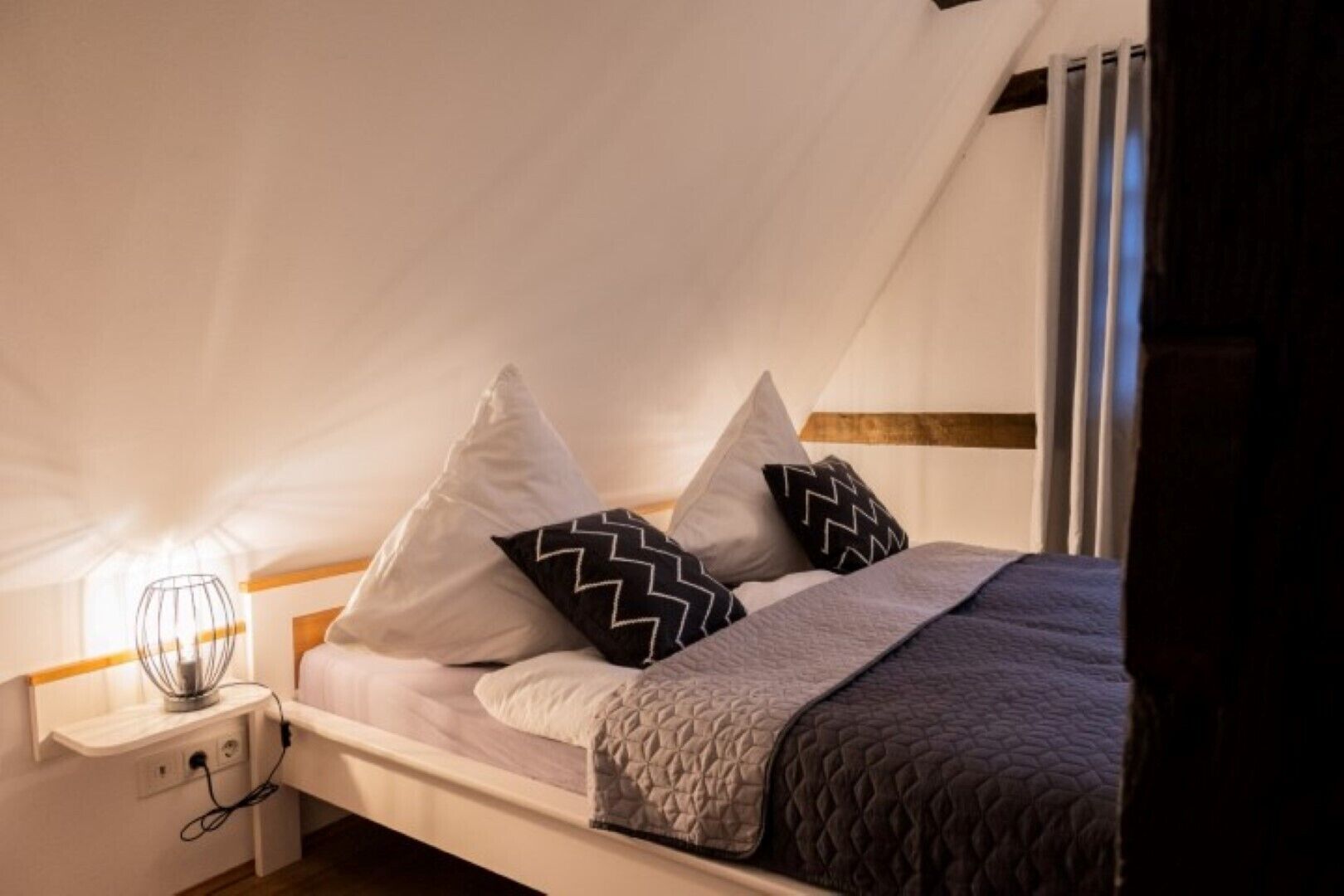 A neatly made bed with a white headboard, two pillows with black zigzag patterns, and a grey quilted blanket.