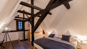 A bedroom with a sloped ceiling, exposed wooden beams, a bed with a dark quilt, and a floor lamp.