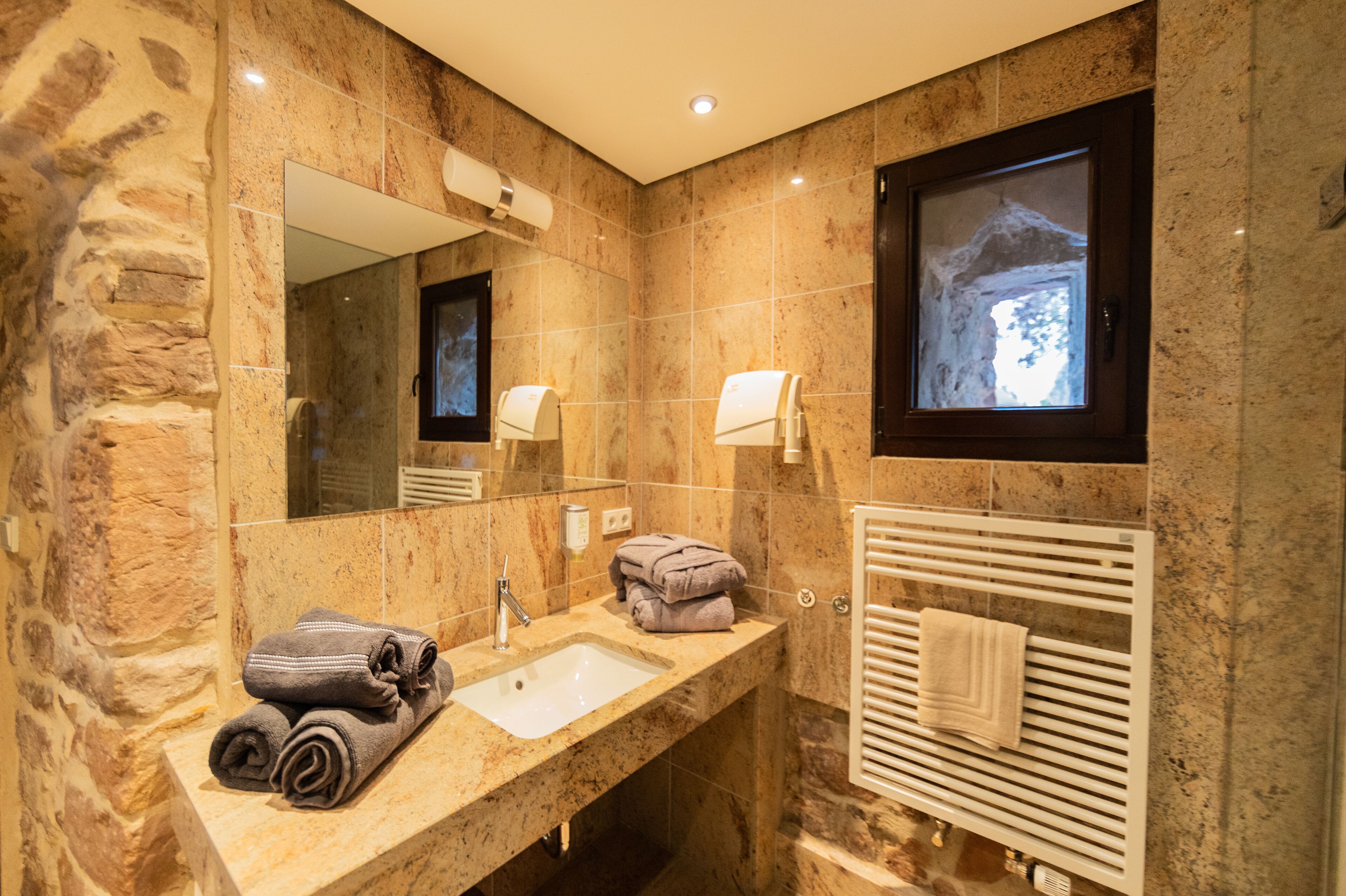 A bathroom with a stone wall, a sink, a mirror, a towel rack, and a window with a view.
