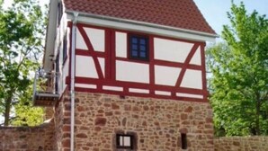 A traditional half-timbered building with a stone facade, a red-tiled roof, and a small entrance with a door. There are potted plants in front of the building.