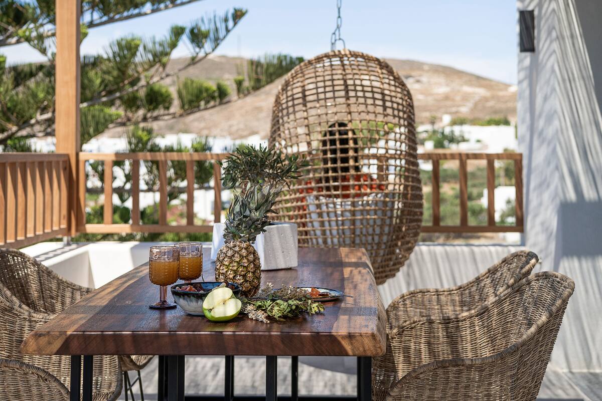 A wooden table with a woven basket, a pineapple, a bowl of fruit, and two glasses of orange juice on a balcony with wicker chairs and a view of a mountainous landscape.