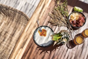 A wooden table with a woven basket, a bowl of fruit, and two glasses of a yellow beverage. - Athina Milos Suites (Milos)