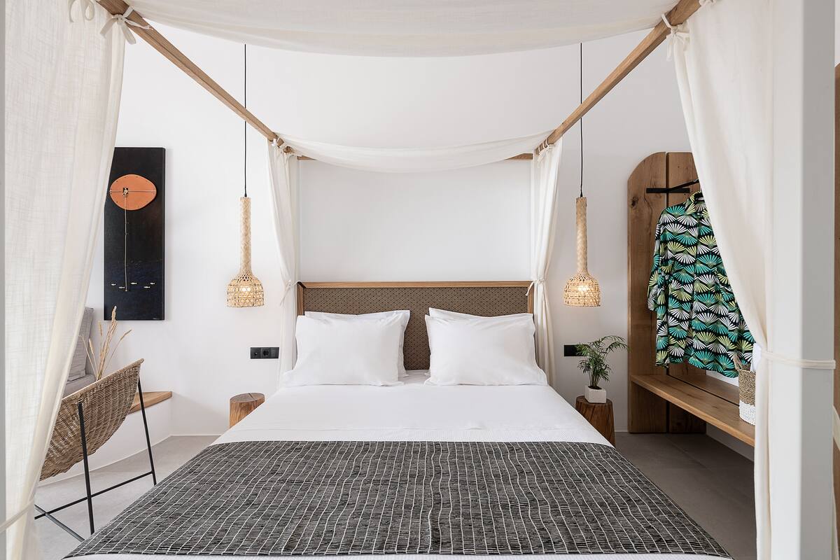 A neatly arranged bedroom with a canopy bed, a woven chair, a bedside table with a plant, and a wall-mounted artwork.