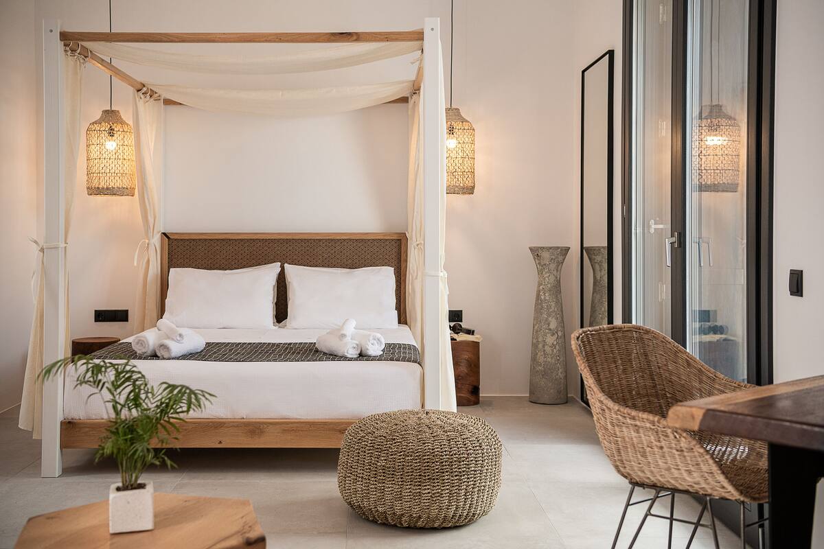 A bedroom with a four-poster bed, a wicker ottoman, a wooden table, and a wicker chair.