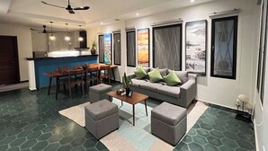 Ein modernes Wohnzimmer mit einer Couch, Hocker, einem Couchtisch und einem Essbereich mit Stühlen und einem Tisch. Es gibt Deckenventilatoren, Kunstwerke an den Wänden und eine Küchenzeile im Hintergrund.