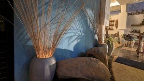 Ein Zimmer mit blauer Wand, einer großen Vase mit hohen Gräsern, einem Stein und einem Essbereich mit Stühlen und Tisch.