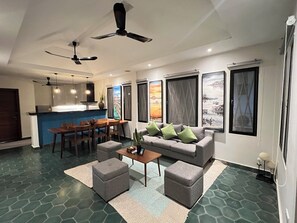 A living room with a sofa, ottomans, a coffee table, and a dining area with chairs and a table. There are ceiling fans, artwork on the walls, and a kitchen in the background. - Home-like Family/Group Stay in Three-Bedroom Suite- Near Pubstreet, Old Market (Krong Siem Reap)
