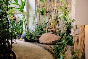 Um jardim interno bem cuidado, com diversas plantas, uma grande rocha como peça central e uma estátua de uma divindade.