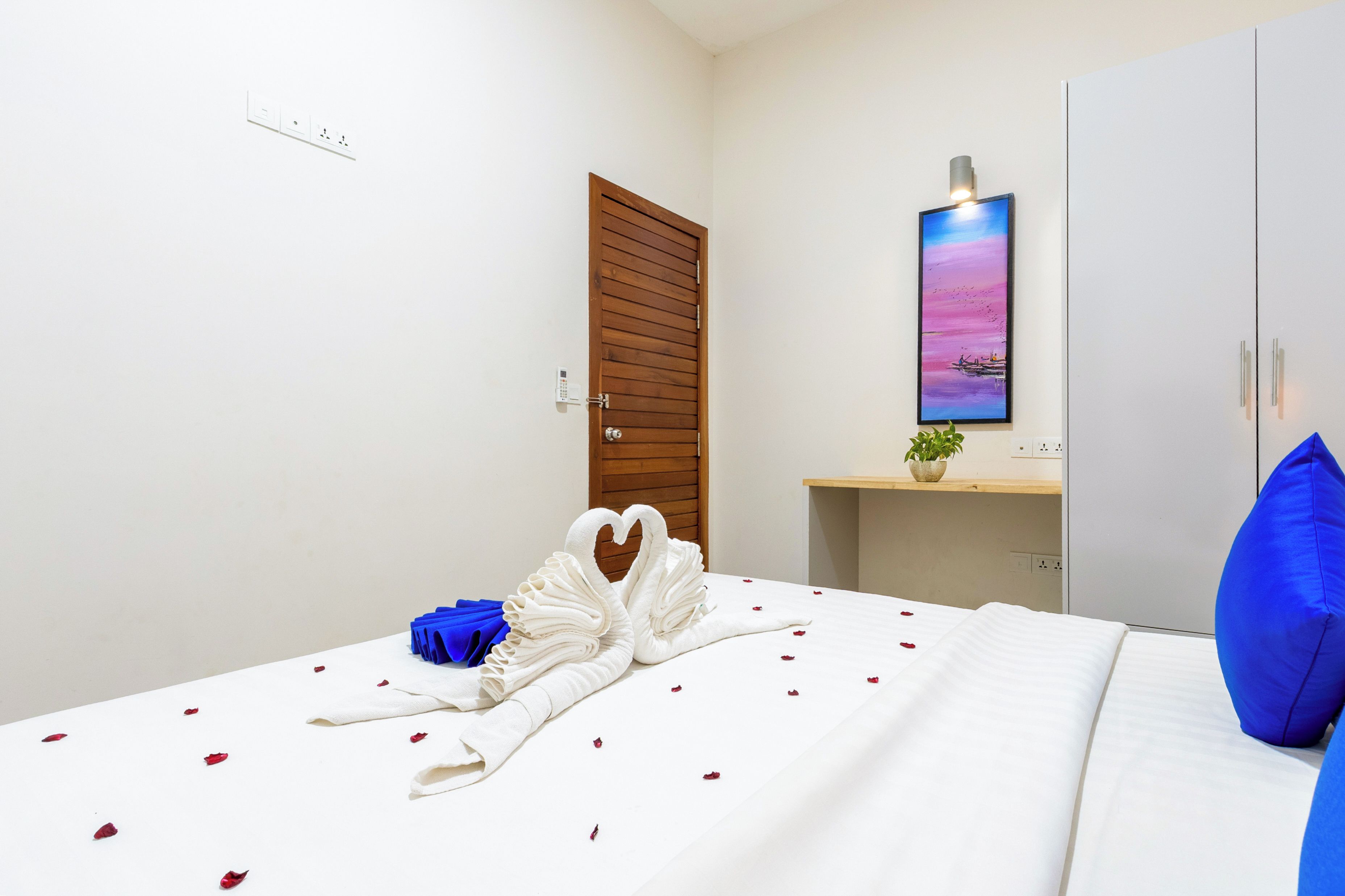 A hotel room with a bed, a wooden door, a painting, a small plant, and a blue pillow.