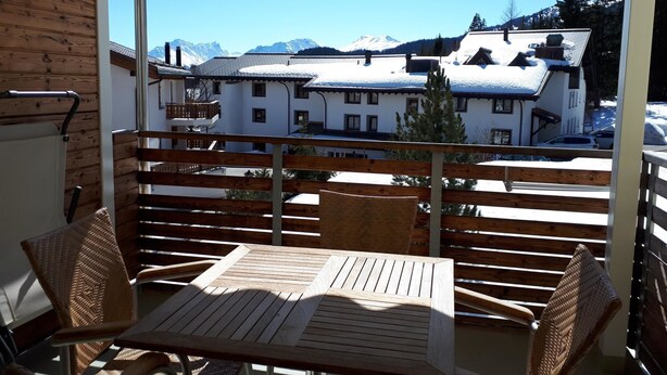 Un balcón con muebles de madera, vista de edificios nevados y montañas al fondo.
