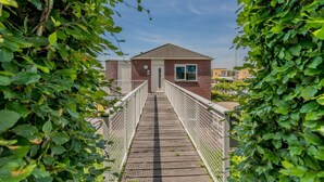 Eine Holzbrücke führt zu einem Backsteinhaus mit einem einzelnen Fenster, umgeben von Grünpflanzen.