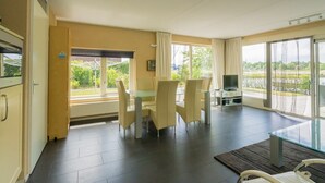 Ein Zimmer mit Esstisch, Stühlen, Fernseher und großen Fenstern mit Blick auf Grünflächen.